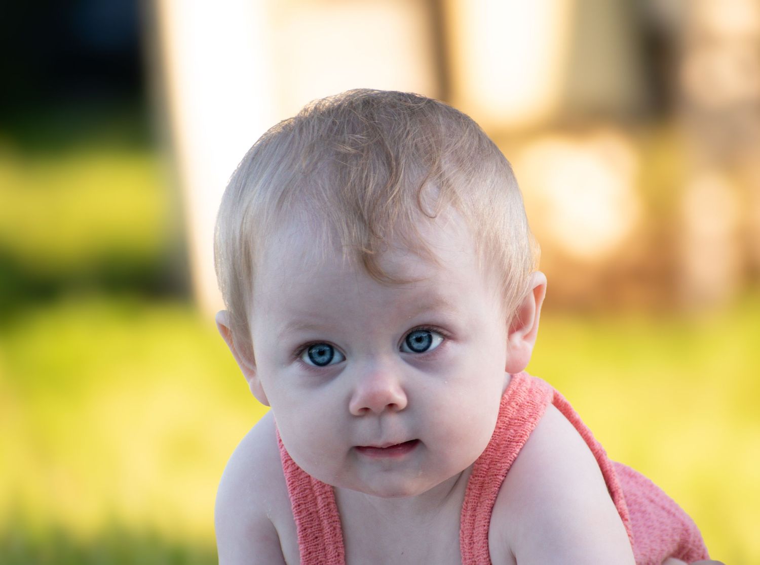 Close-up portrait of a blue-eyed baby in a pink romper against a soft green bokeh background.