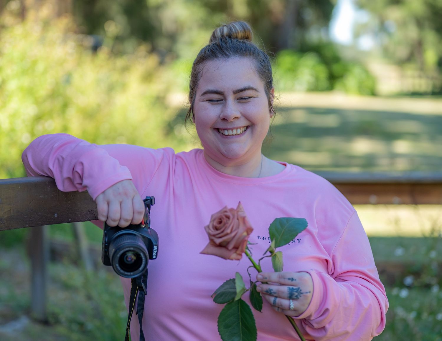 Woman in pink sweatshirt smiling, holding a rose and camera, leaning on a wooden fence outdoors.