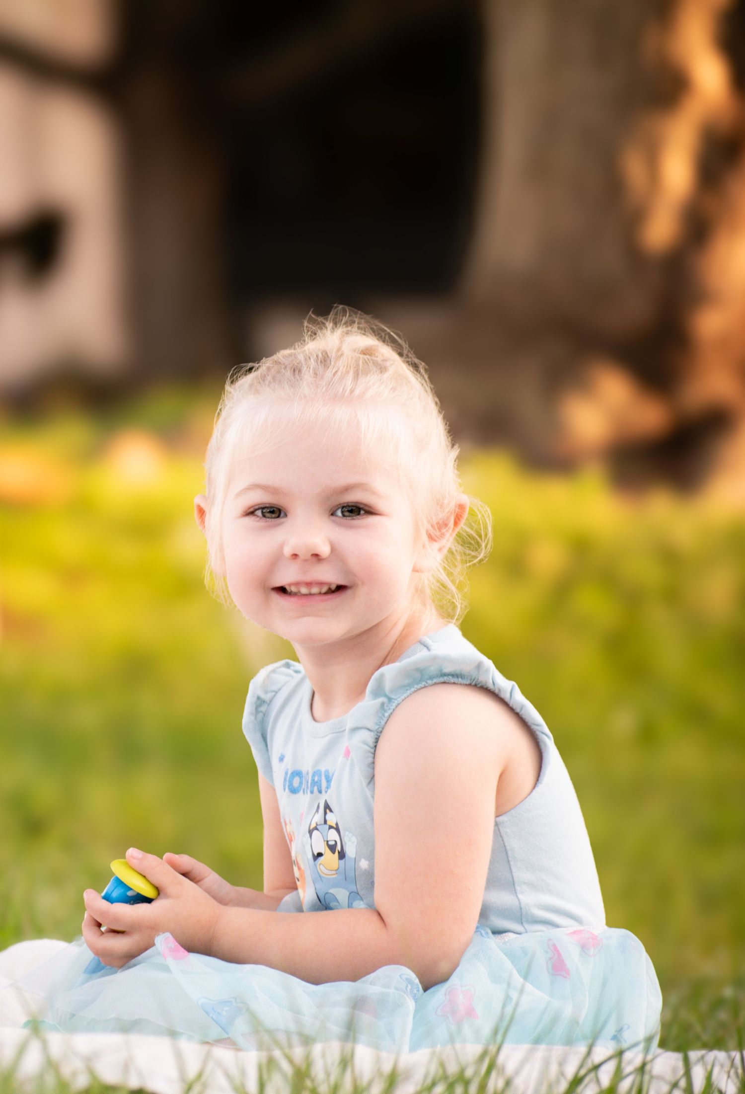 A smiling blonde toddler girl in a blue Bluey dress sits on grass holding a colorful toy.