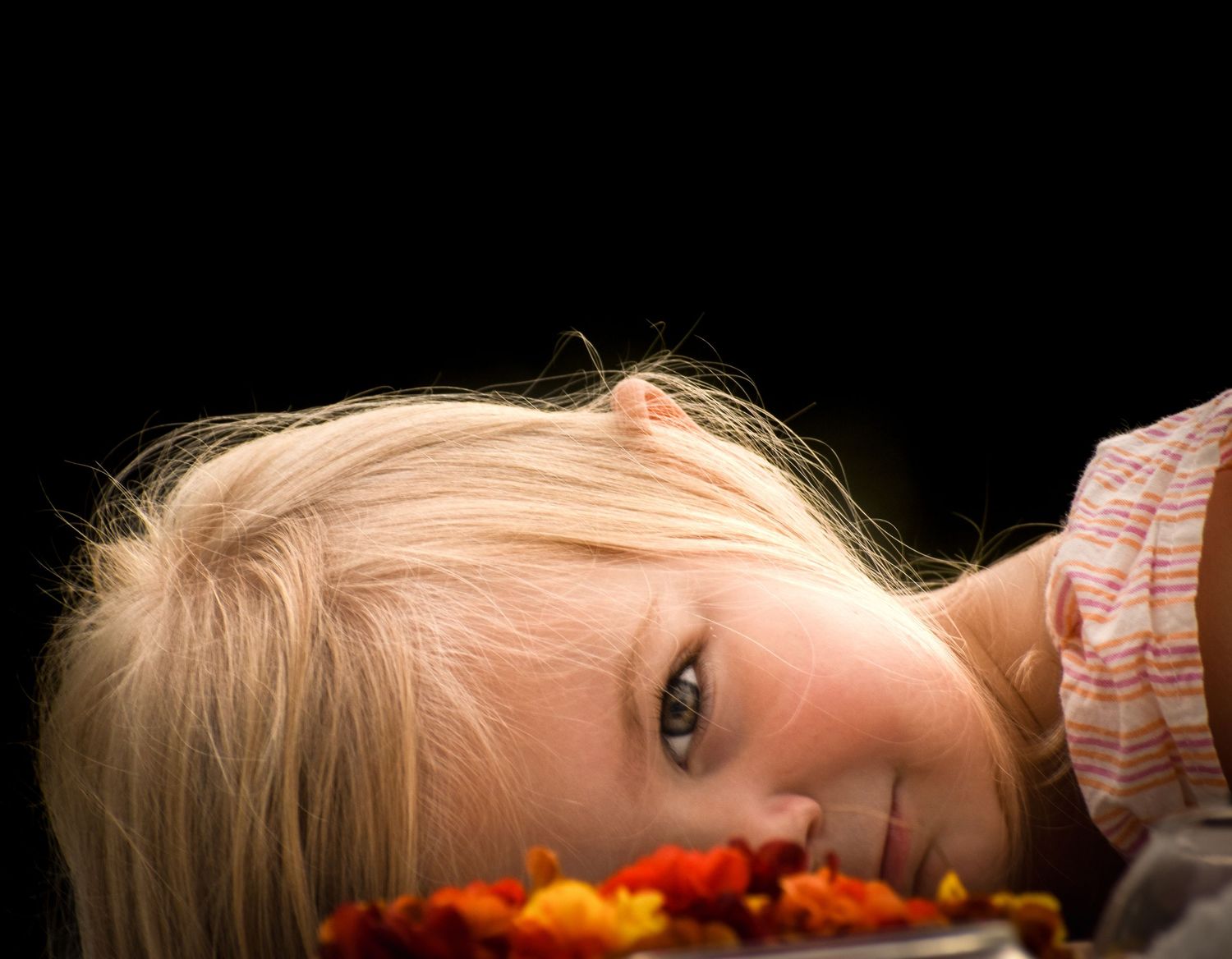 Close-up of blonde girl lying down with colorful flowers against a black background.