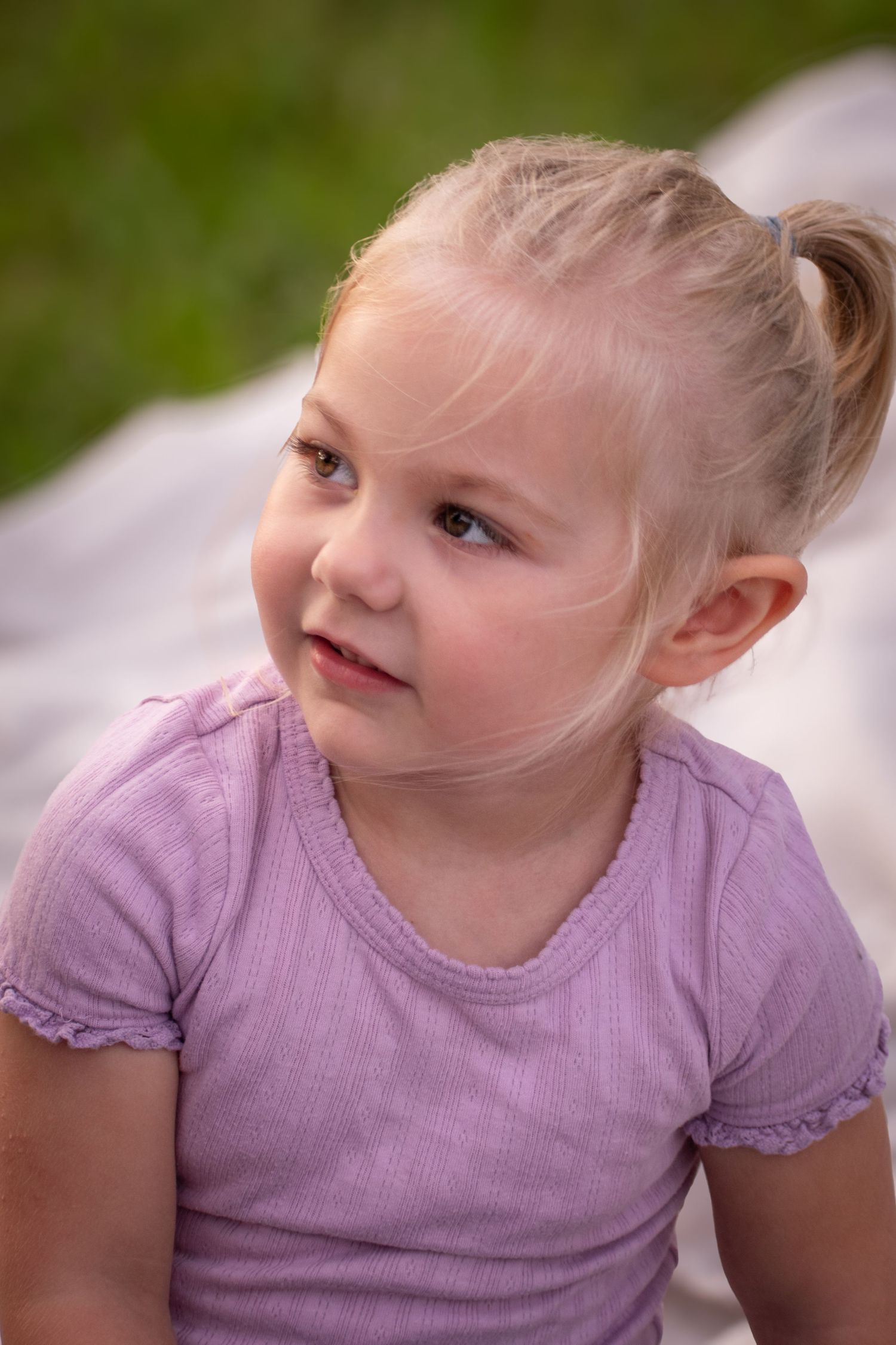 Cute blonde girl with pigtail in lavender shirt sitting outdoors on blanket looking sideways.