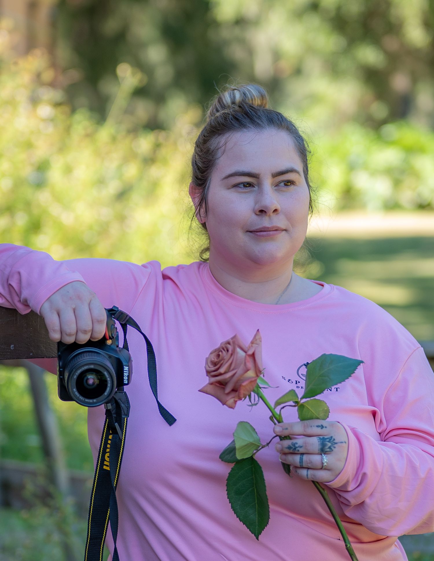 Woman in pink sweatshirt holding a rose and camera outdoors in a lush green garden setting.