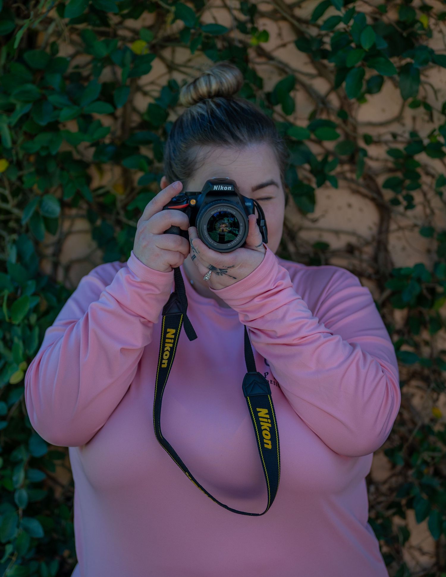 A woman in a pink long-sleeve shirt photographs with a Nikon camera in front of a green ivy-covered wall.