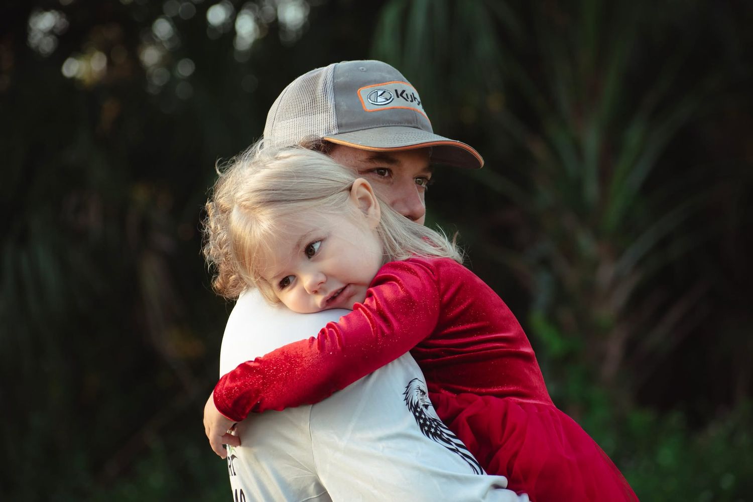 Blonde toddler in red shirt hugging an adult wearing a gray cap outdoors near palm trees.