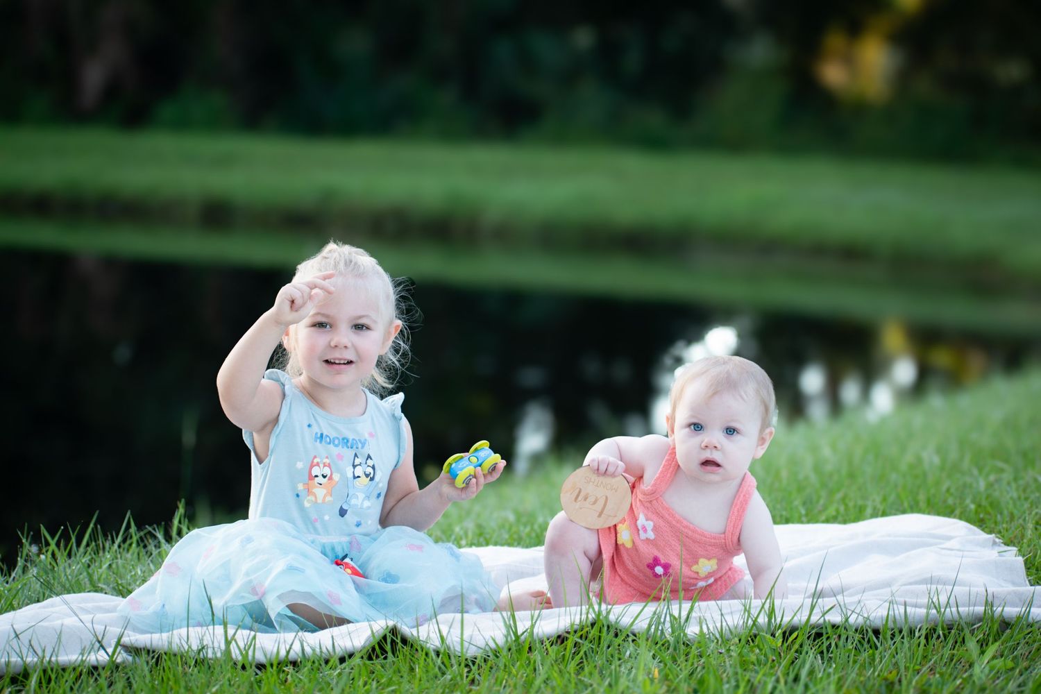 Two young sisters play on a white blanket in green grass near a pond on a sunny day.