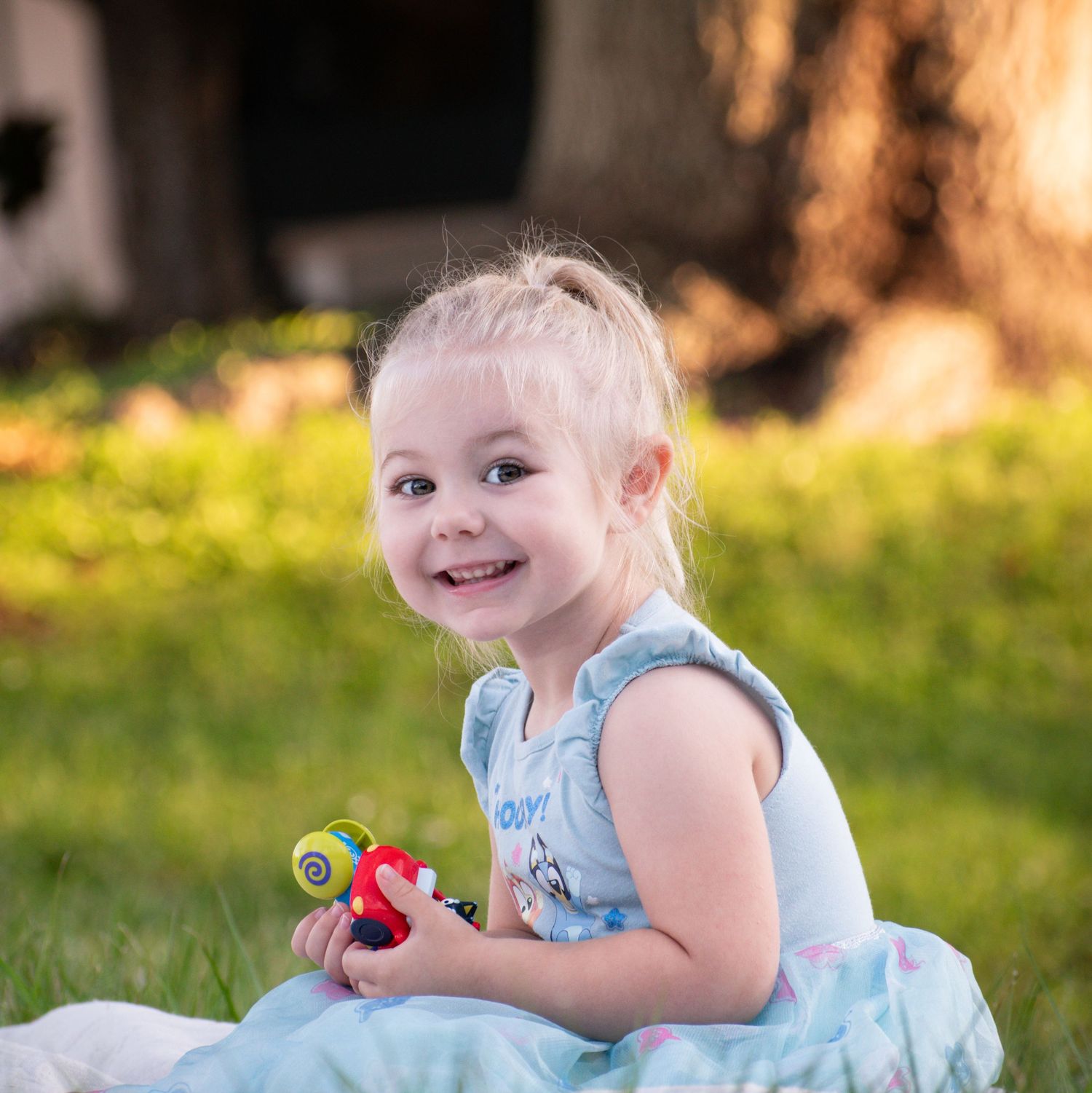 Smiling blonde toddler girl in blue dress sitting on grass holding colorful toy in sunny outdoor setting.