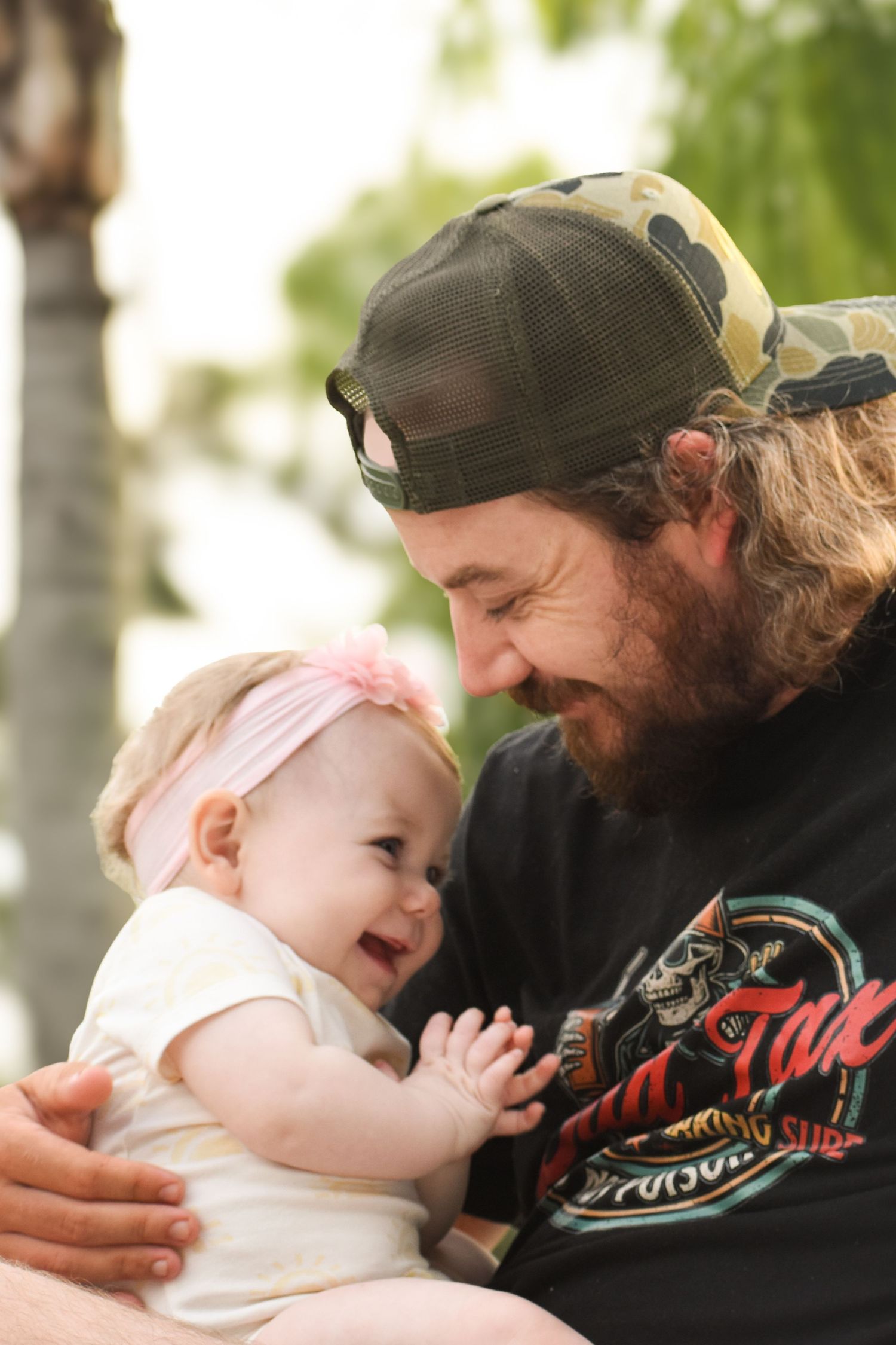 Bearded dad in camo cap smiling at giggling baby girl wearing pink headband outdoors.