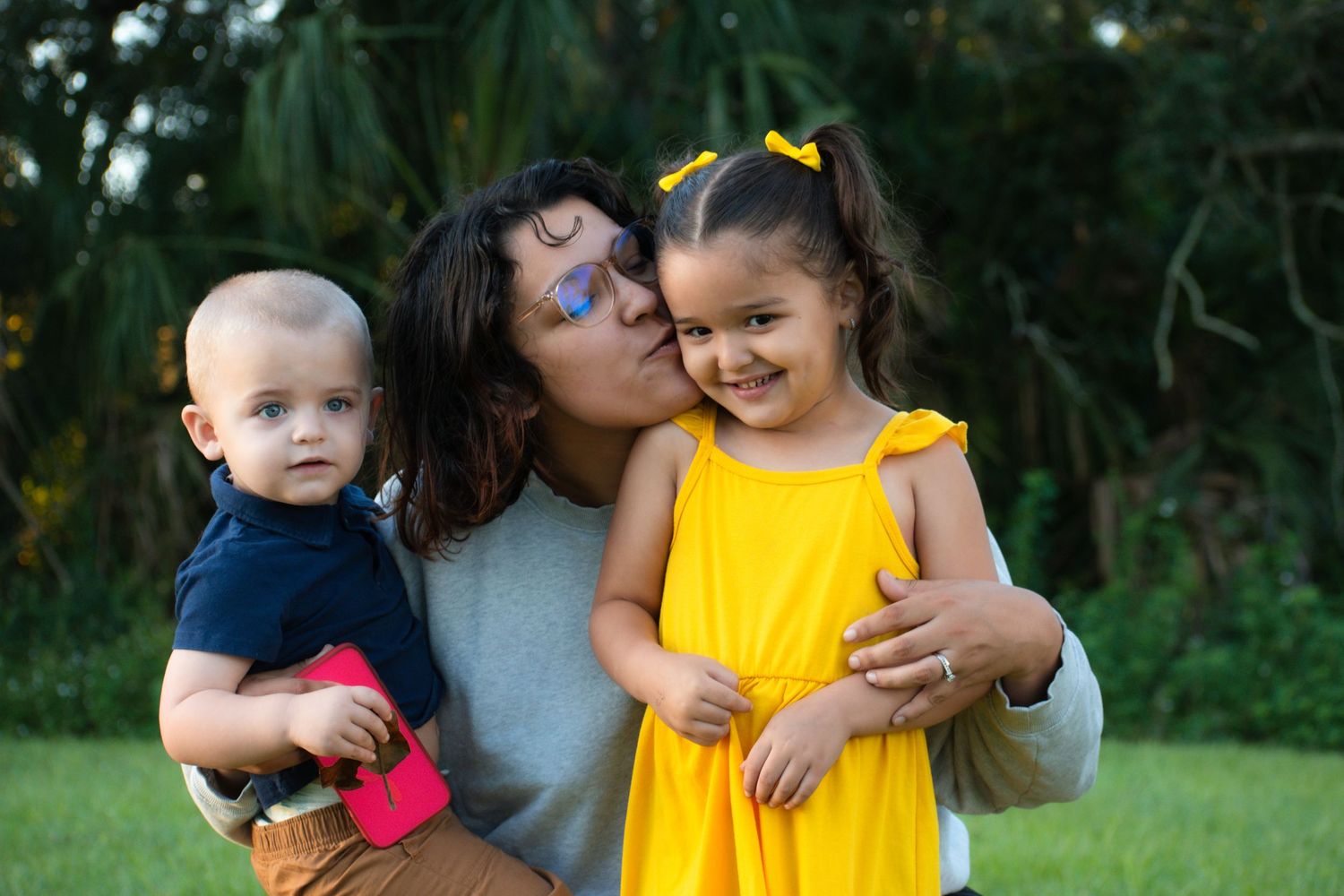 Mother kissing young girl in yellow dress while holding baby boy in outdoor park setting.