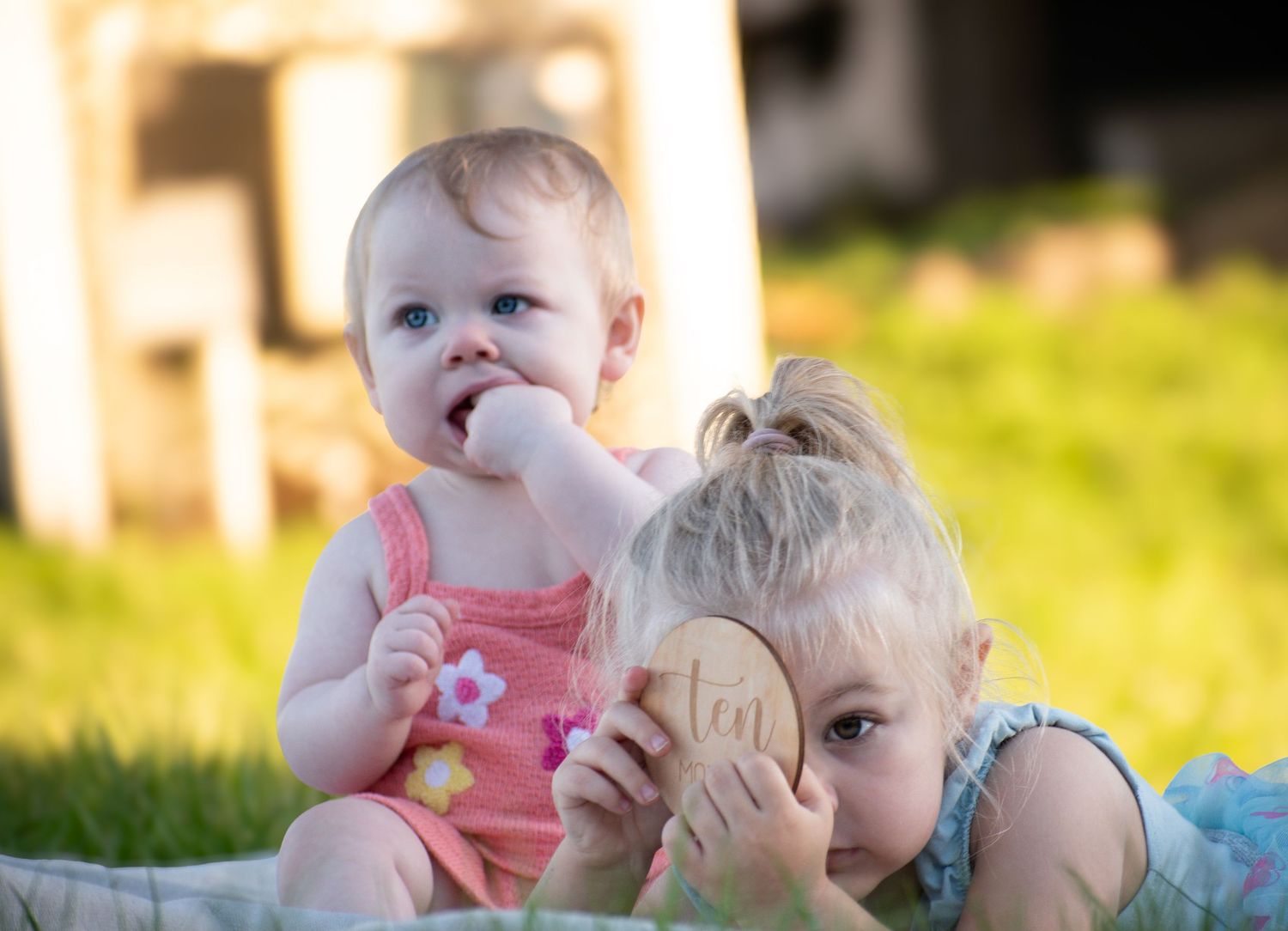Two babies sit on grass outdoors, one in pink romper chewing fingers while older toddler holds a wooden toy.