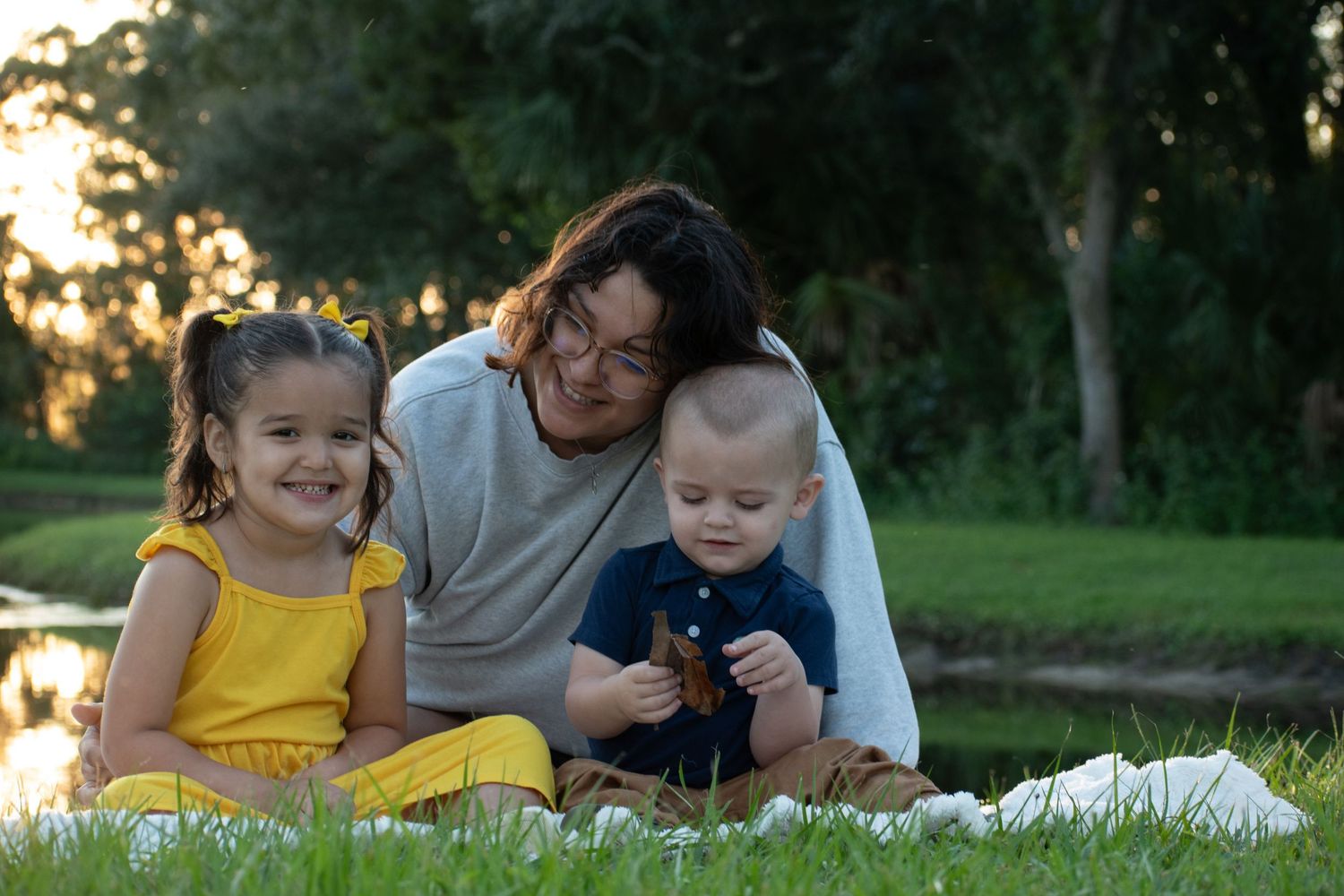 Mother with two young children sitting on grass blanket by pond during golden sunset.