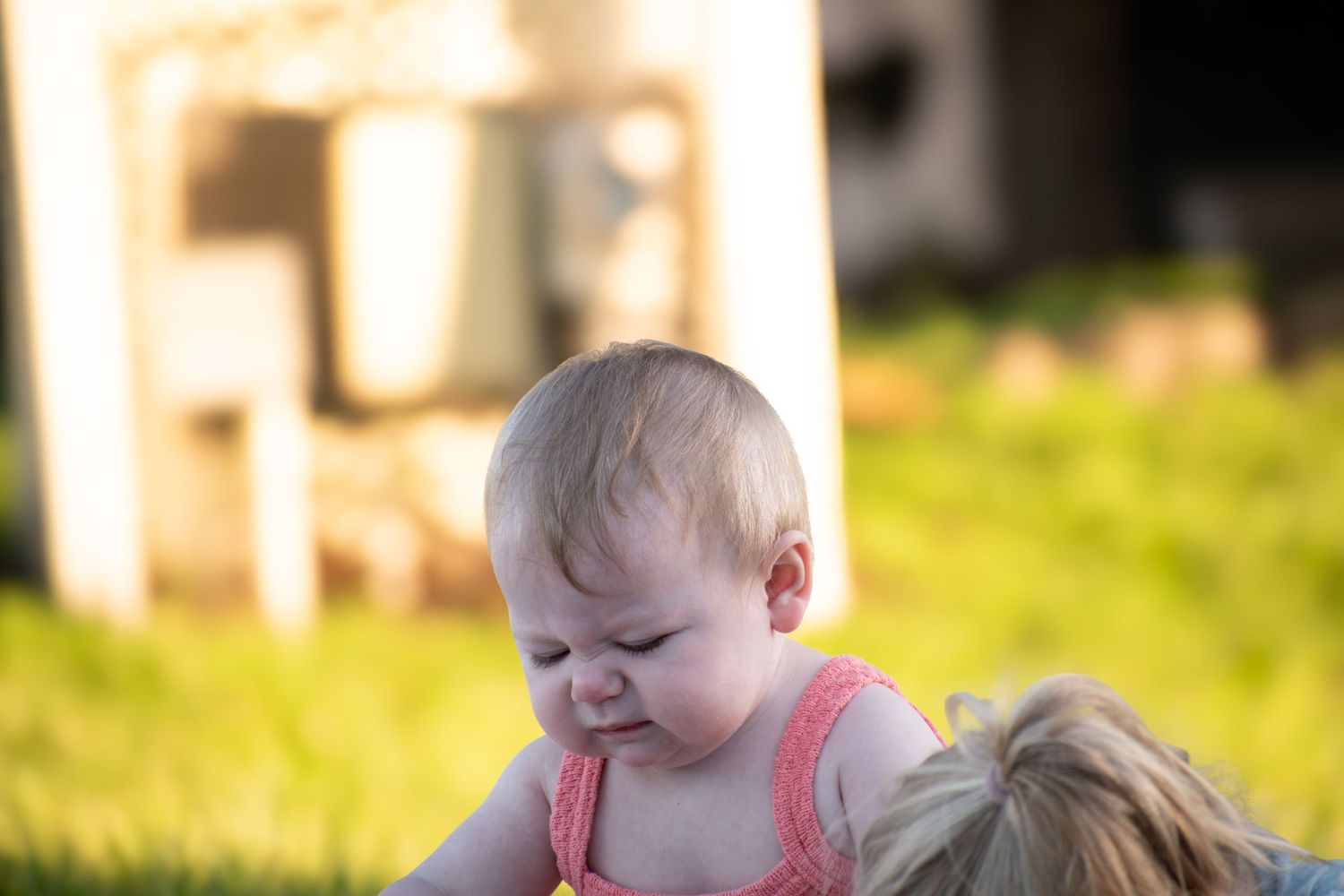 Baby in pink romper makes a scrunchy face while being held on green grass in golden sunlight.