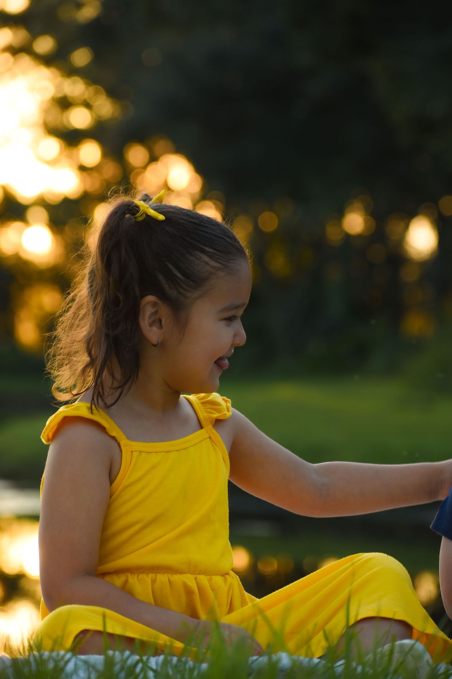 Happy little girl in yellow dress sitting on grass laughing during golden sunset outdoors.
