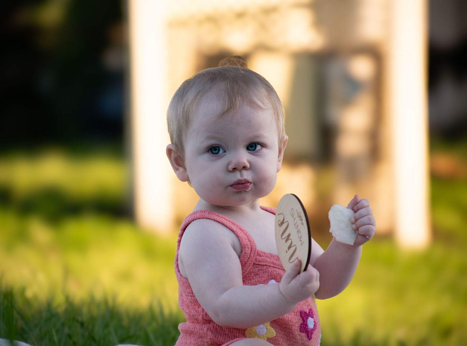 Cute blonde baby girl in pink dress sitting on grass holding a wooden keepsake disc outdoors.