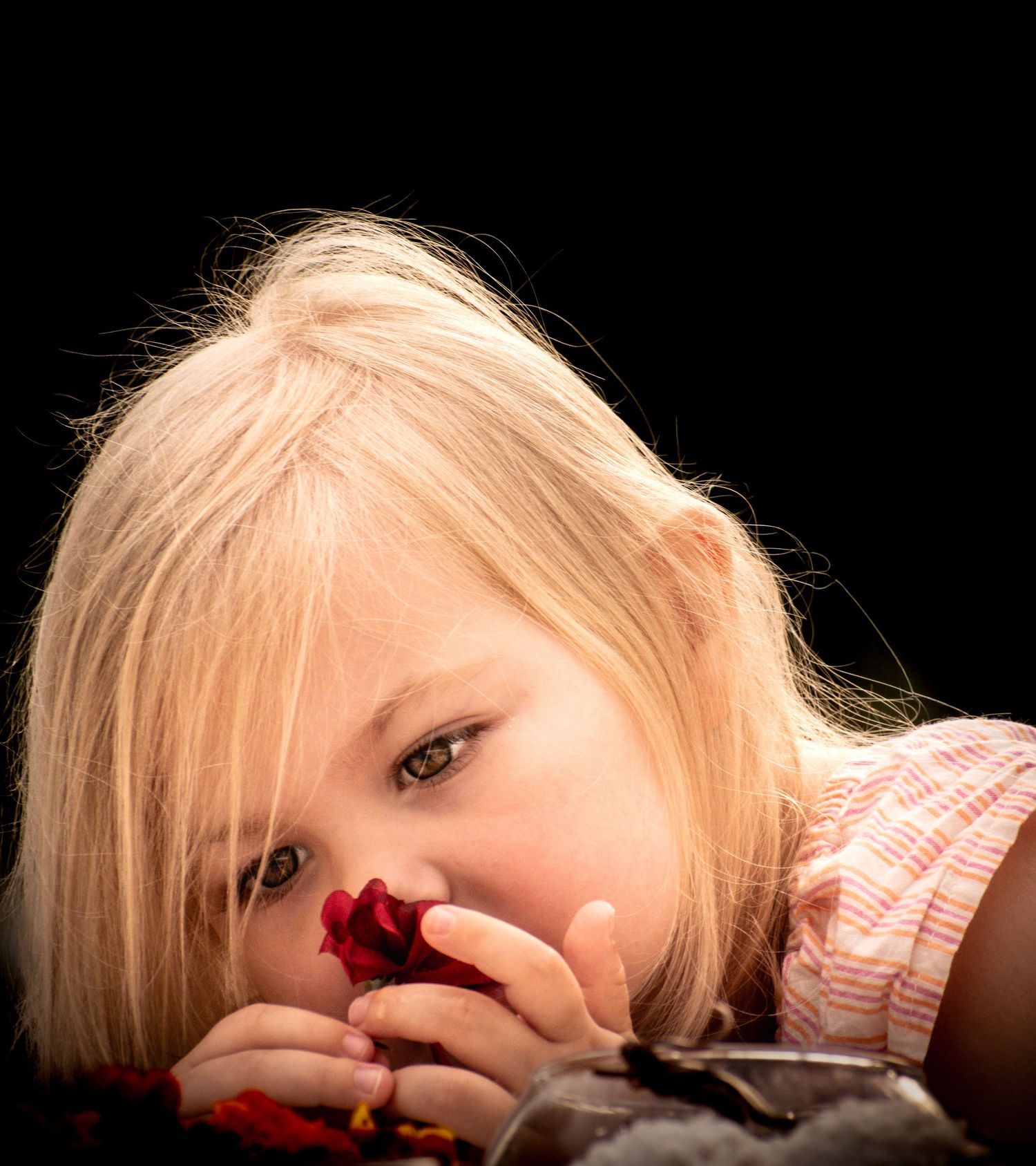 Close-up of blonde girl lying down holding a red rose against dark black background.