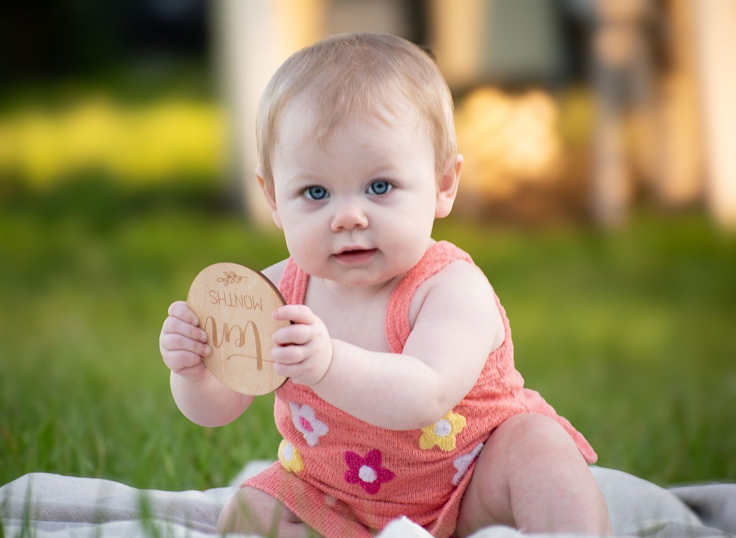 An adorable baby girl in a pink floral romper holds up a wooden milestone disc on green grass.