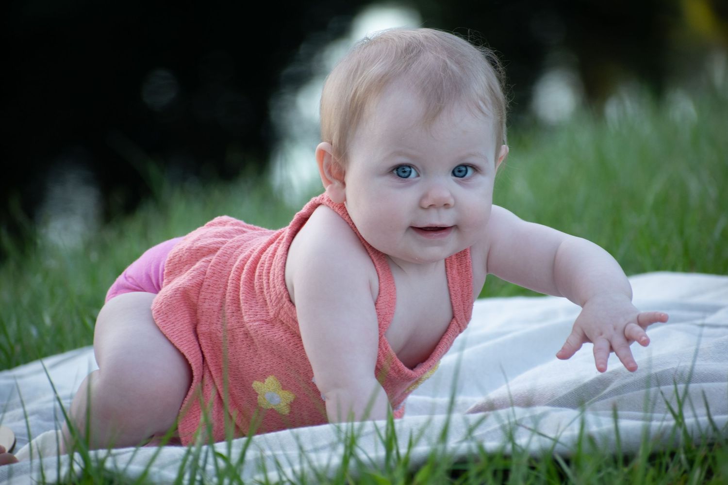 A blue-eyed baby girl in a pink romper does tummy time on a white blanket in green grass.
