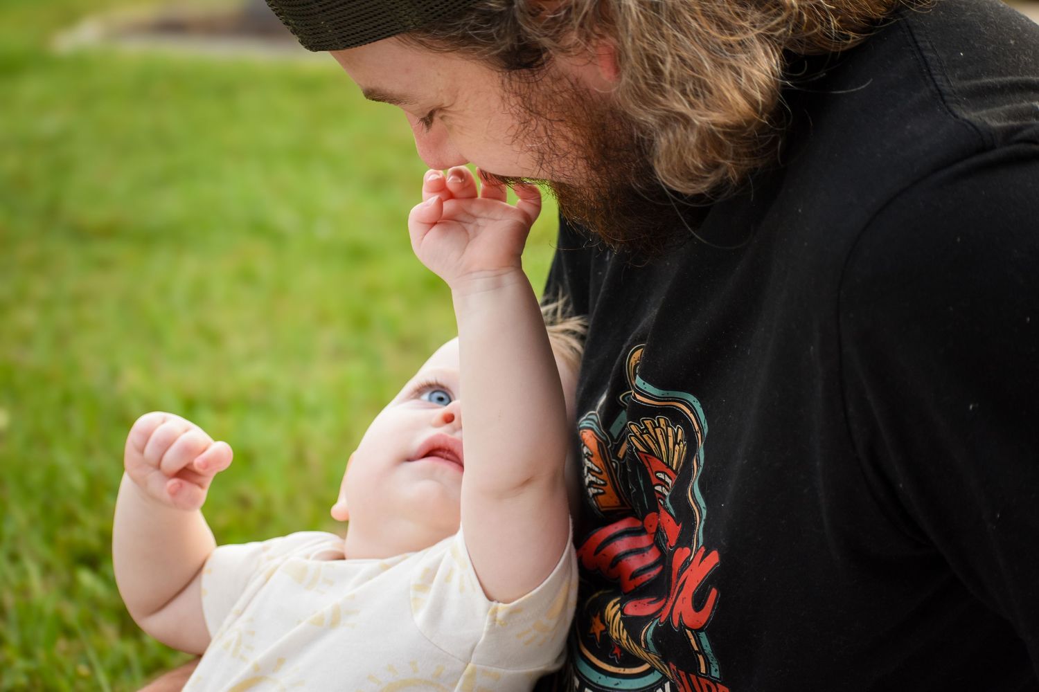 Loving bearded father gently kissing hand of curious baby lying in his arms outdoors.