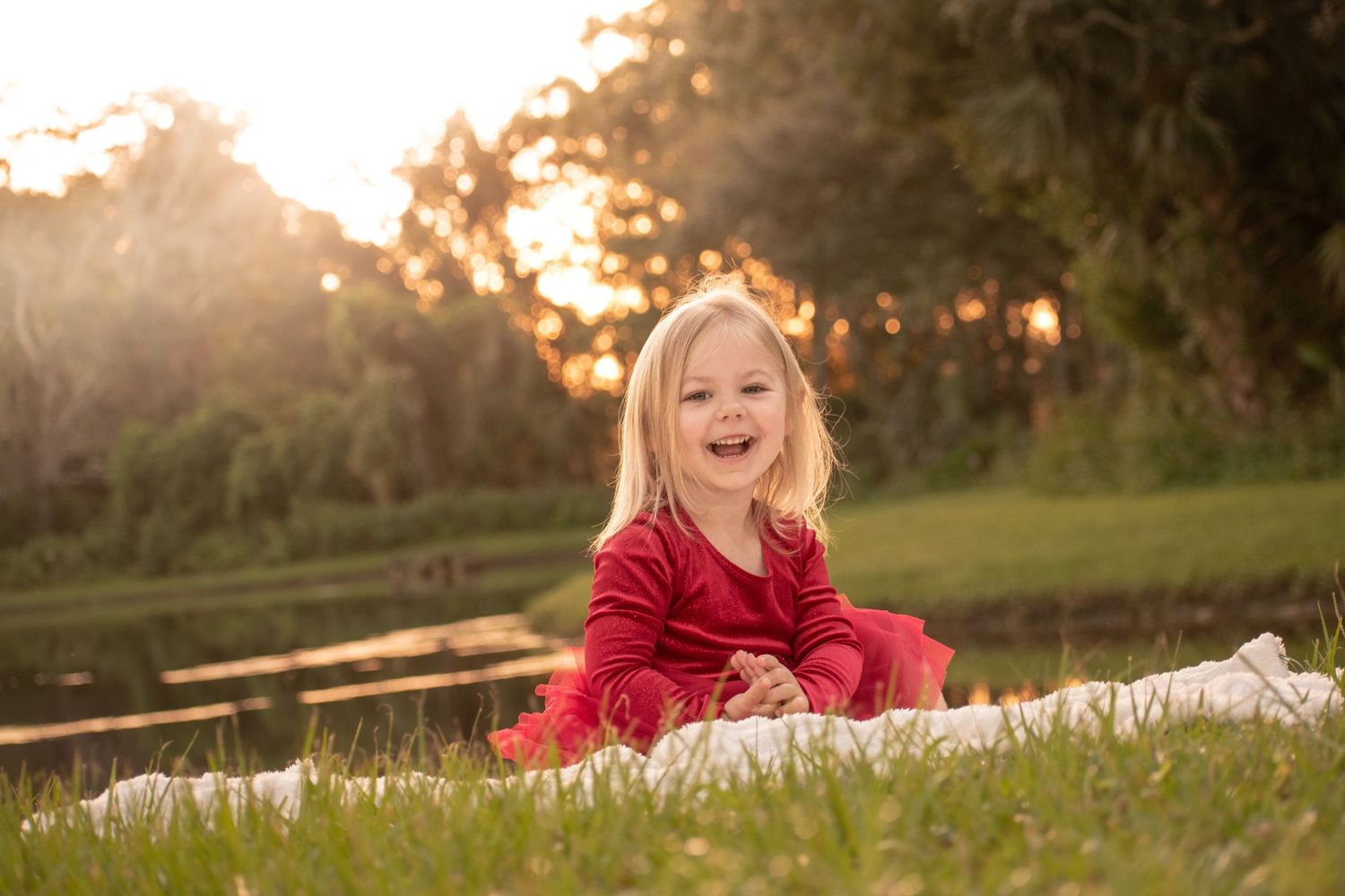 Smiling blonde toddler girl in red dress sitting on grass during golden hour sunset in park.