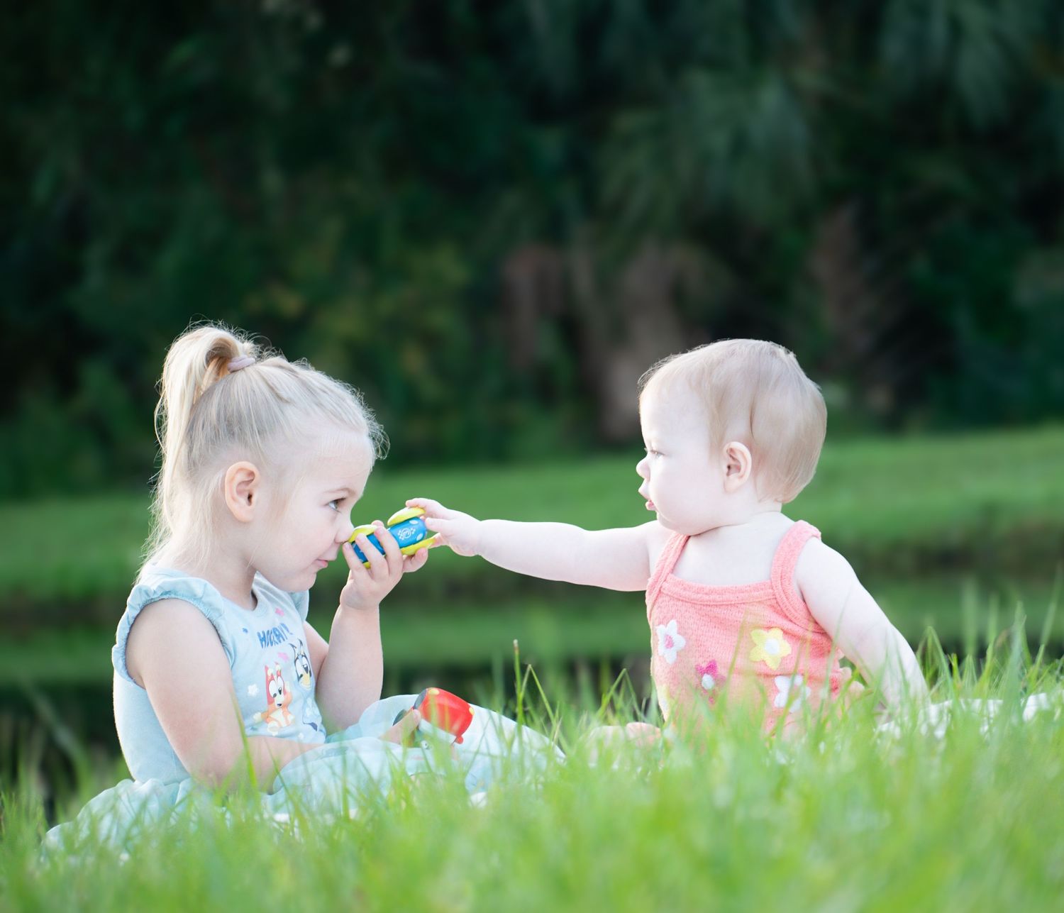 Two young sisters play together on green grass, with the baby reaching out to share a toy.