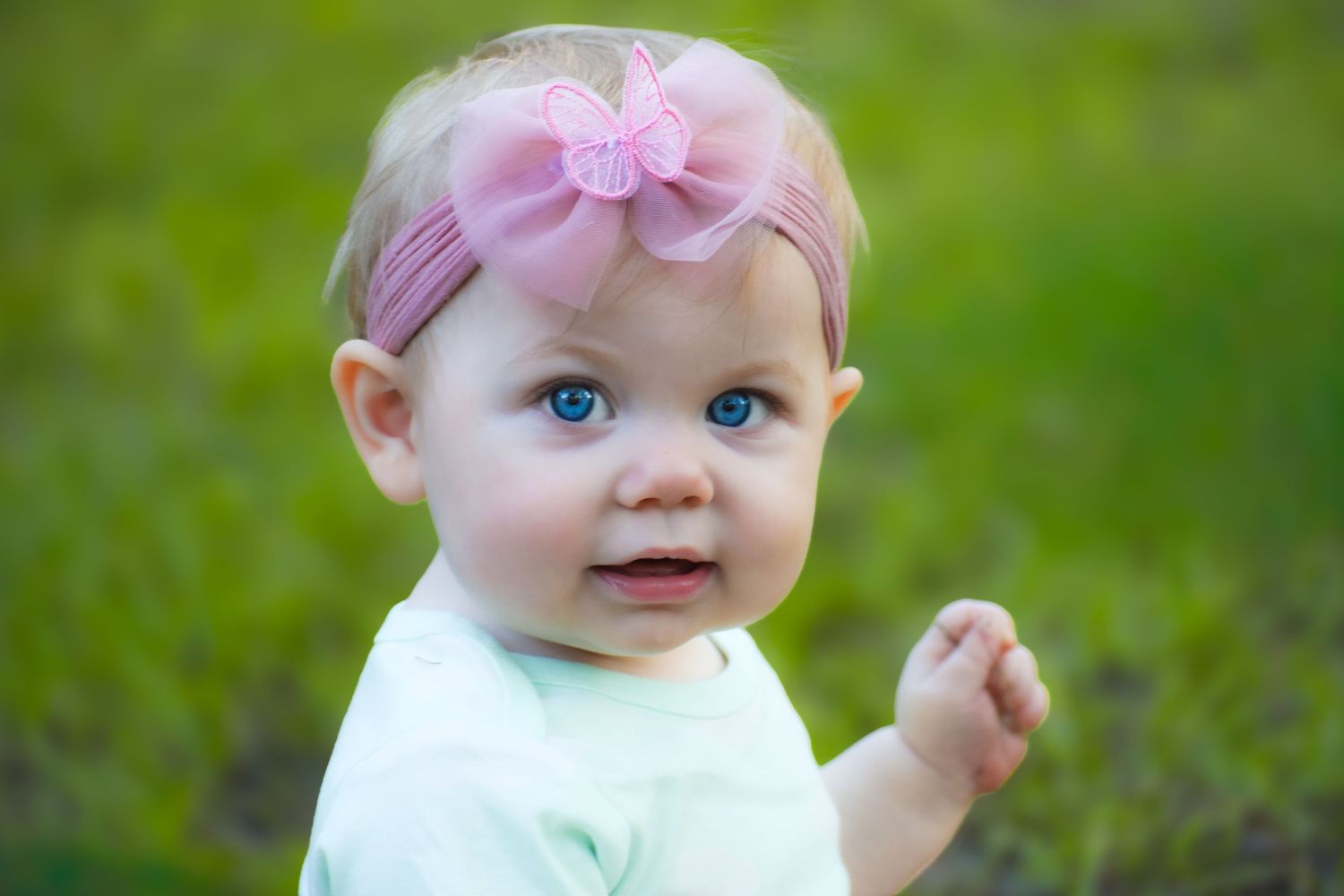 Cute baby girl with blue eyes and pink bow headband looking back over shoulder on grass.