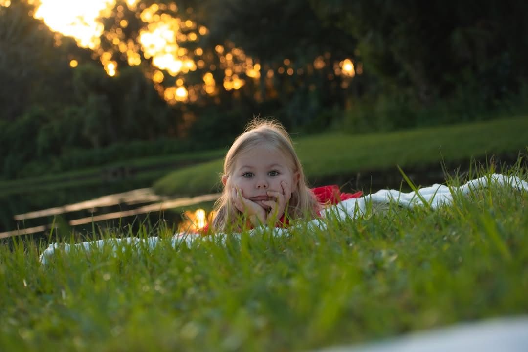 Young blonde girl lying in lush green grass during golden hour sunset in a park.
