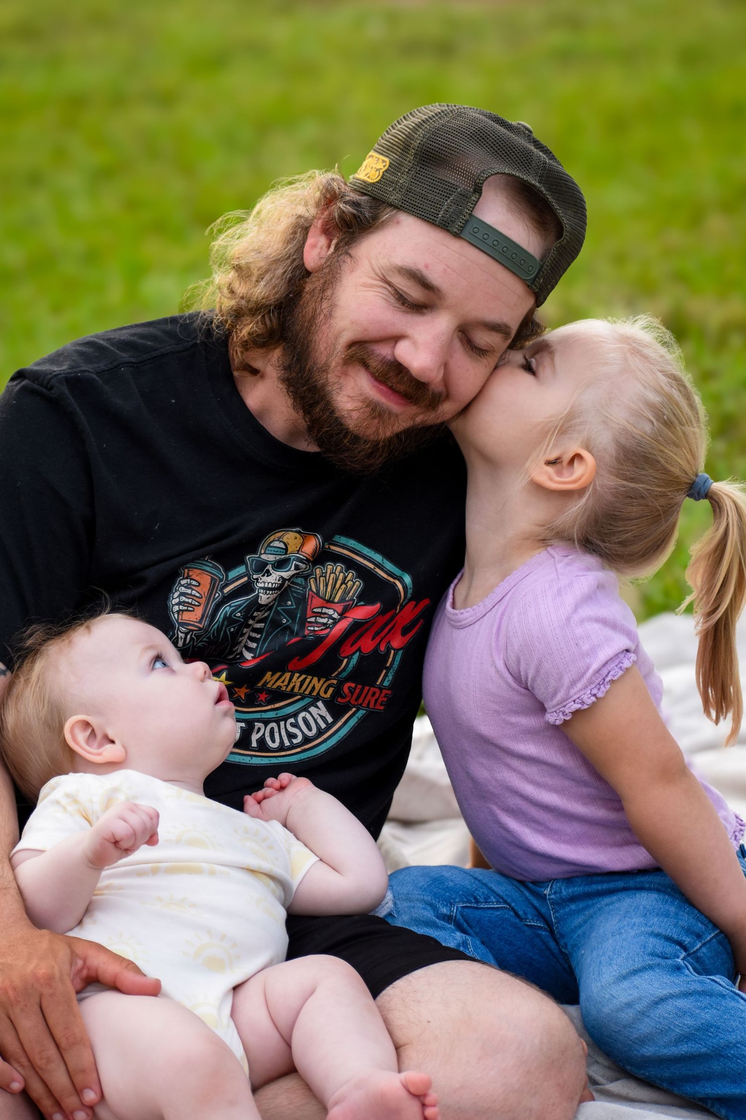 A bearded dad smiles as his young daughter kisses his cheek while holding a baby outdoors.