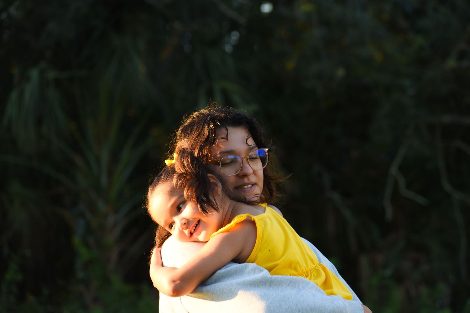 Woman carrying a young girl in a yellow dress outdoors with lush green trees in background.
