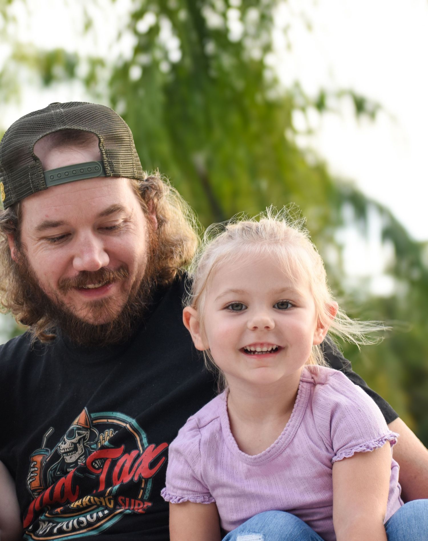 Smiling bearded father in black t-shirt posing with happy blonde toddler daughter outdoors.