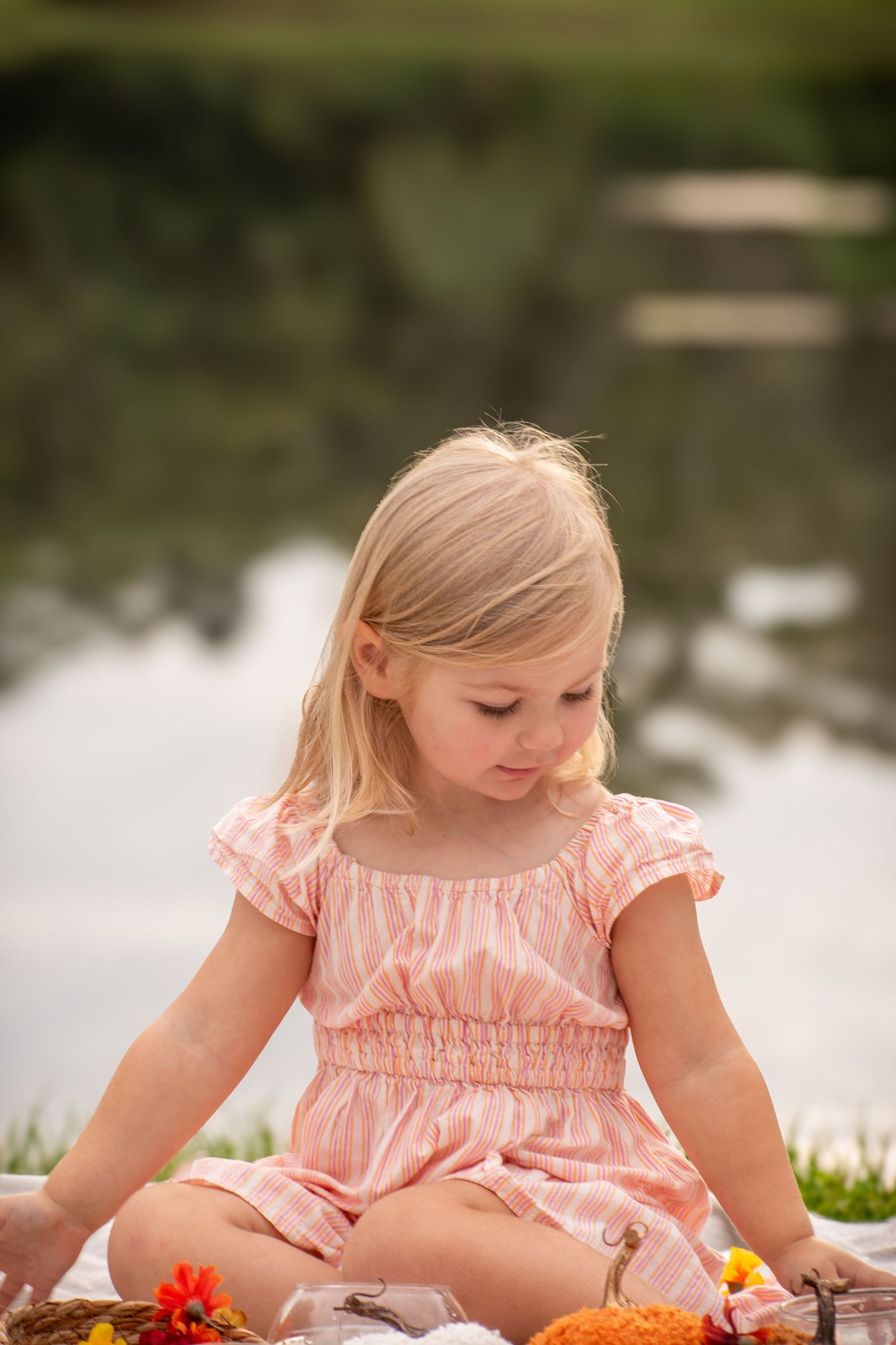 Blonde toddler girl in pink dress sits cross-legged on blanket near a reflective pond outdoors.