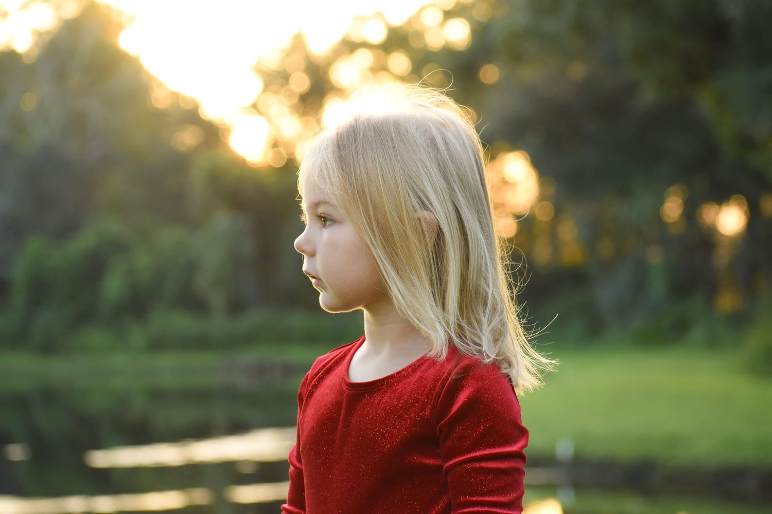 Side profile of blonde little girl in red dress standing near pond in sunlit park.