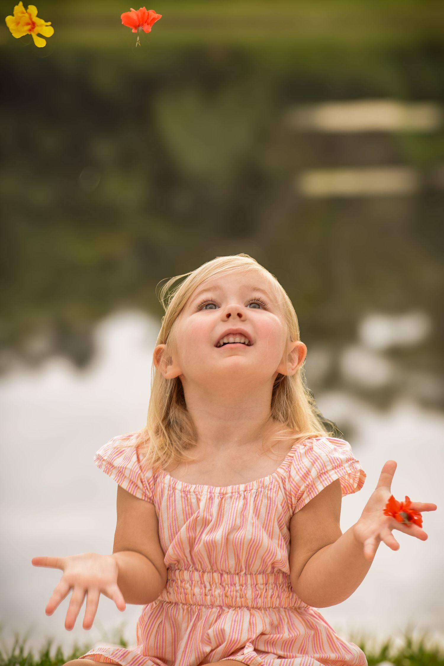 Happy blonde little girl in pink dress tossing colorful flowers in the air by a pond.