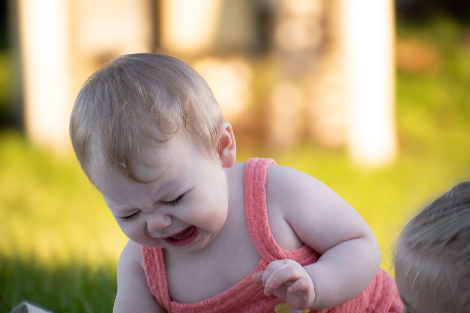 Crying baby in pink sleeveless romper sits on green grass in warm sunlight outdoors.