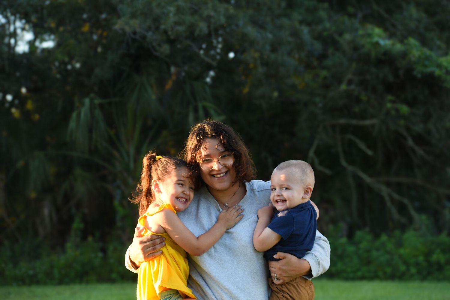Happy mother hugging two young children, girl in yellow dress and baby boy, in sunny park.