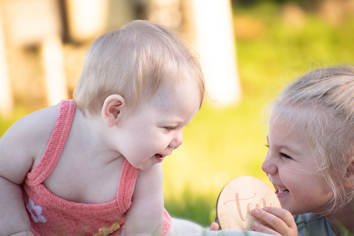 Two young children share a joyful moment outdoors, smiling at each other over a round wooden toy.