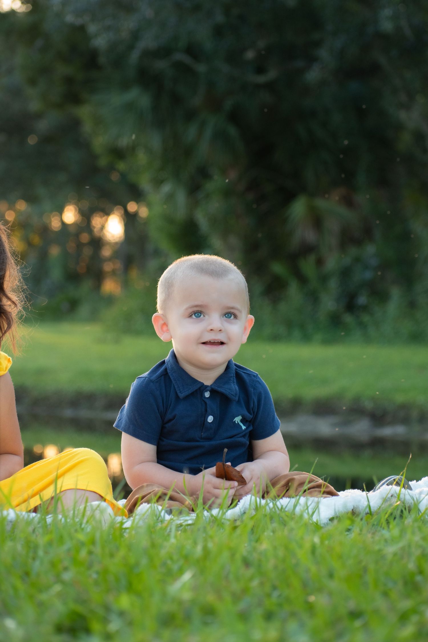 Adorable baby boy in navy polo shirt sitting on white blanket on green grass outdoors.
