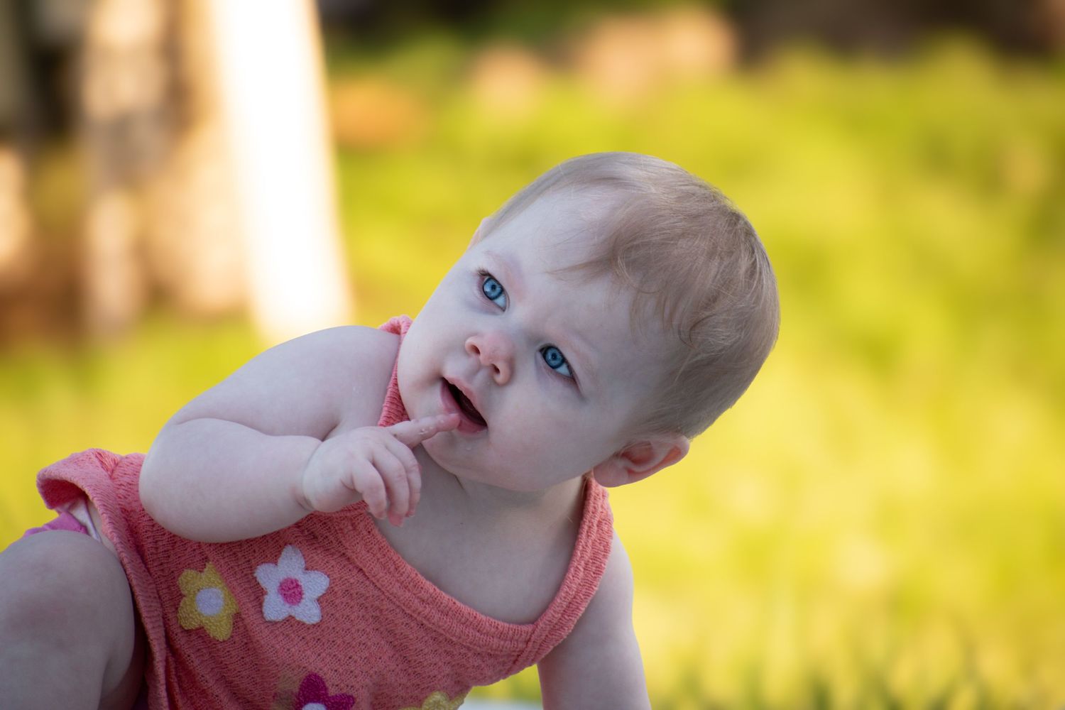 Bright-eyed blonde baby girl in pink floral dress smiling with finger in mouth on green grass.