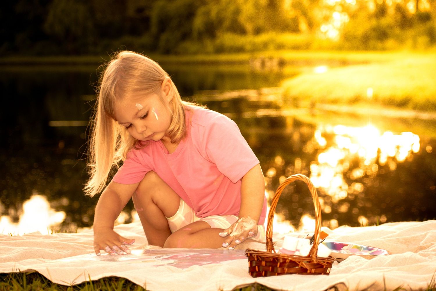 Young blonde girl in pink shirt playing on blanket near pond during golden sunset.