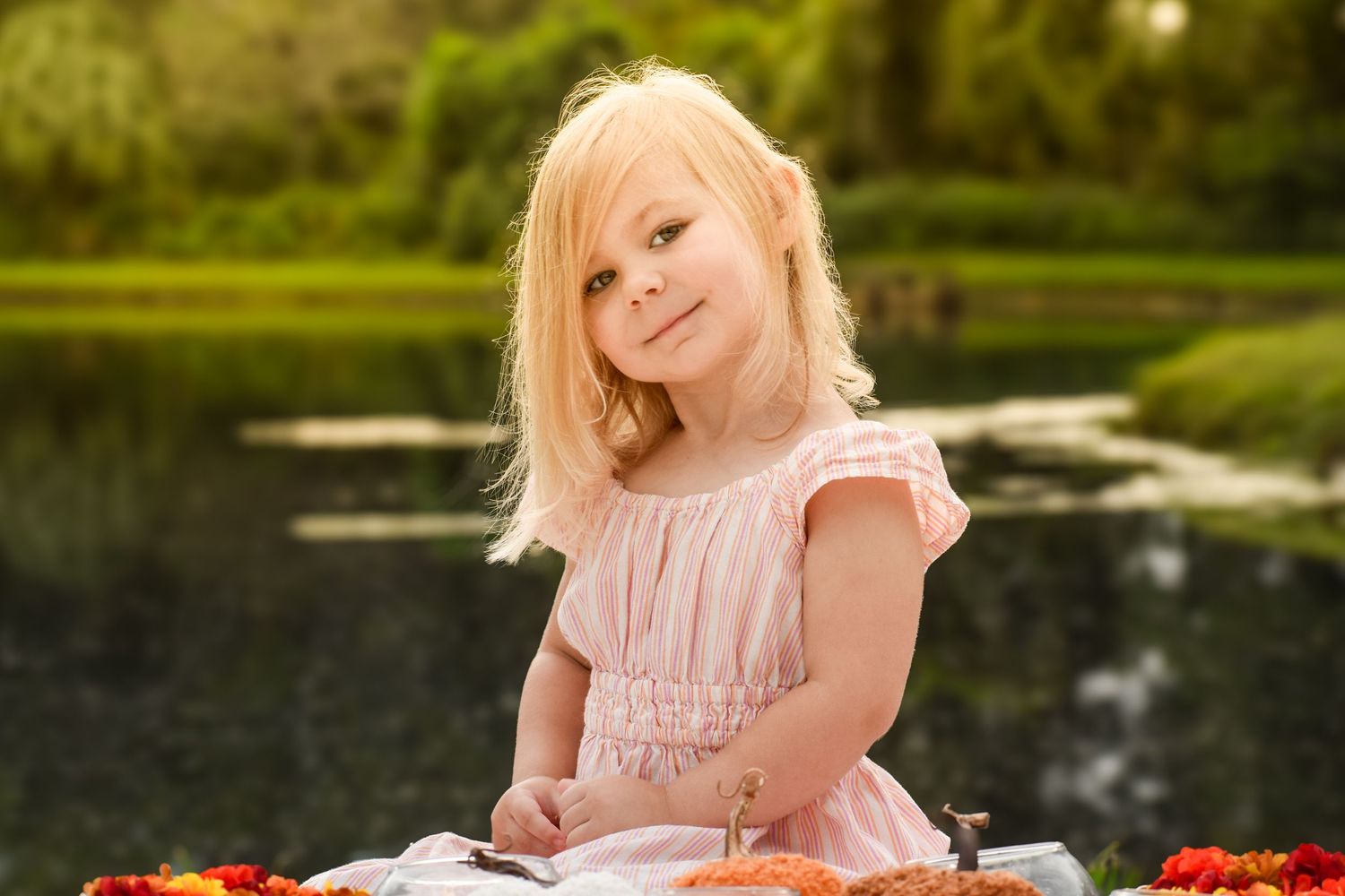 Sweet blonde girl in pink dress sitting at picnic blanket by pond smiling at camera.