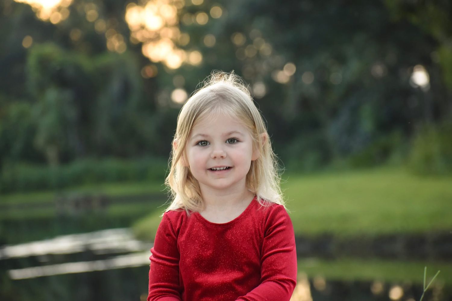 Smiling blonde toddler girl in red long sleeve shirt sitting outdoors near a pond at golden hour.