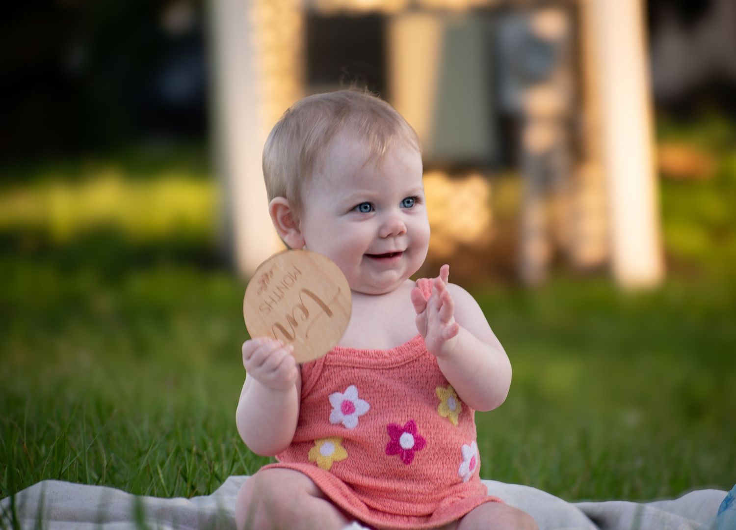 A happy baby girl laughs while holding a wooden milestone disc sitting on grass in sunlight.