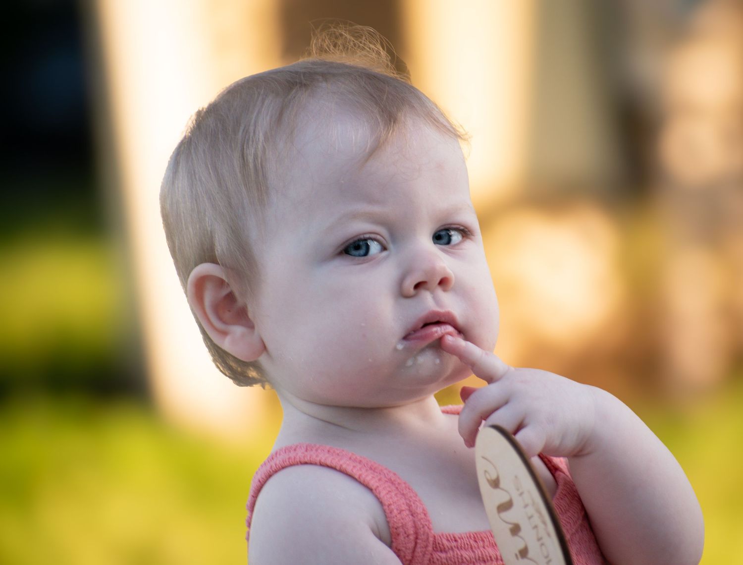 Close-up of a cute baby girl in a pink top holding a wooden teether outdoors.