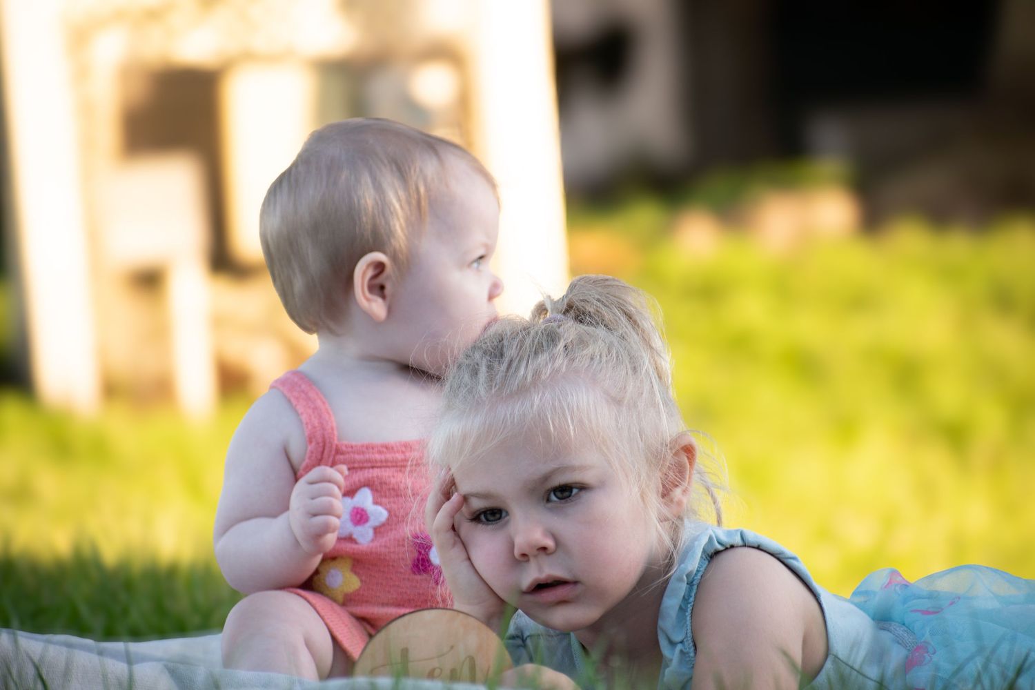 Two young siblings sit together on a blanket in a sunny garden, baby in pink and toddler in blue.