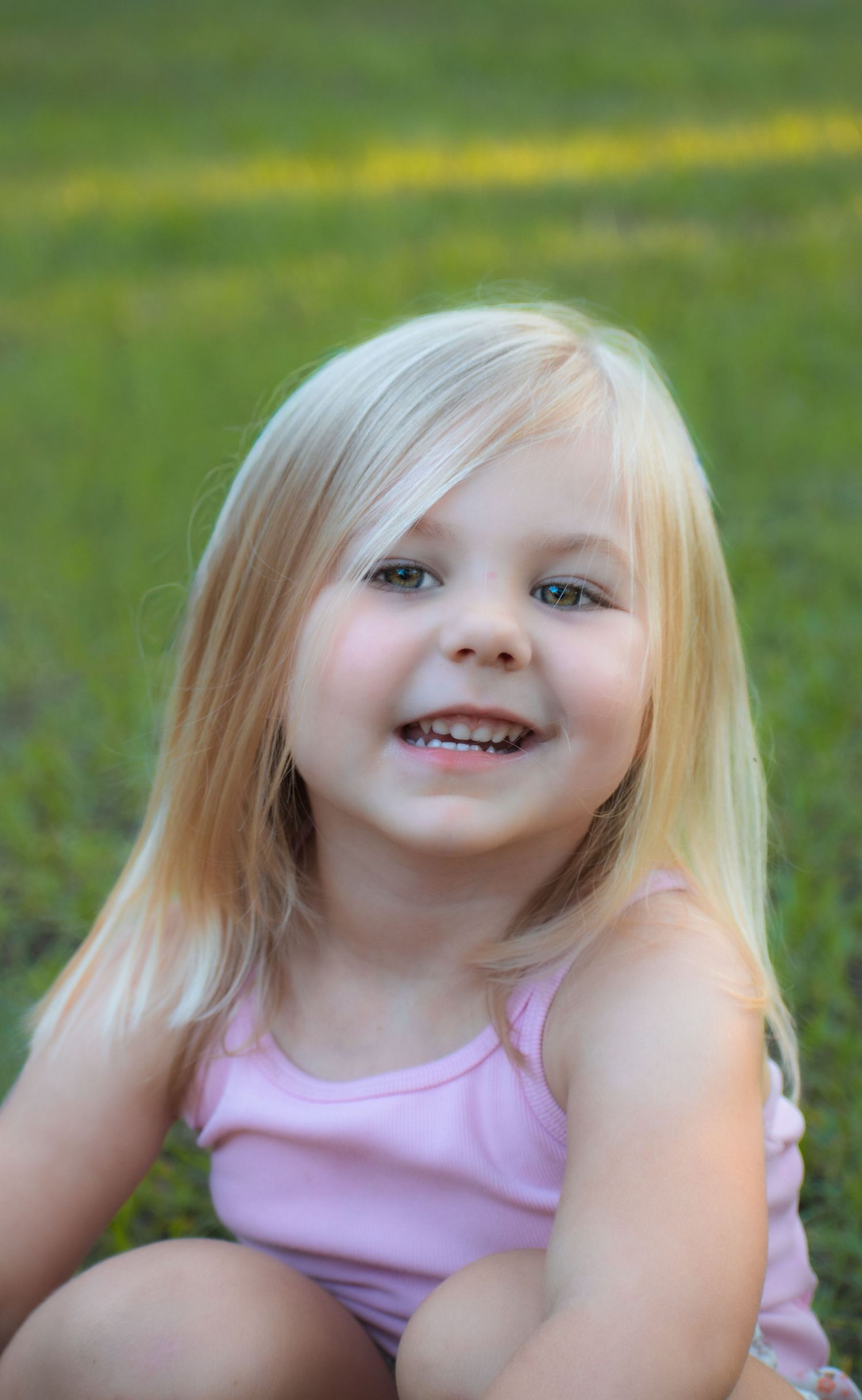 Adorable blonde toddler girl with blue eyes smiling while sitting on green grass outdoors.