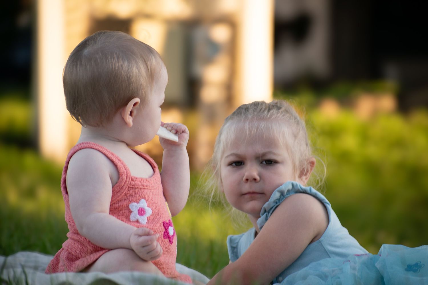 Two little blonde toddler girls sitting together on a blanket in a sunny outdoor setting.