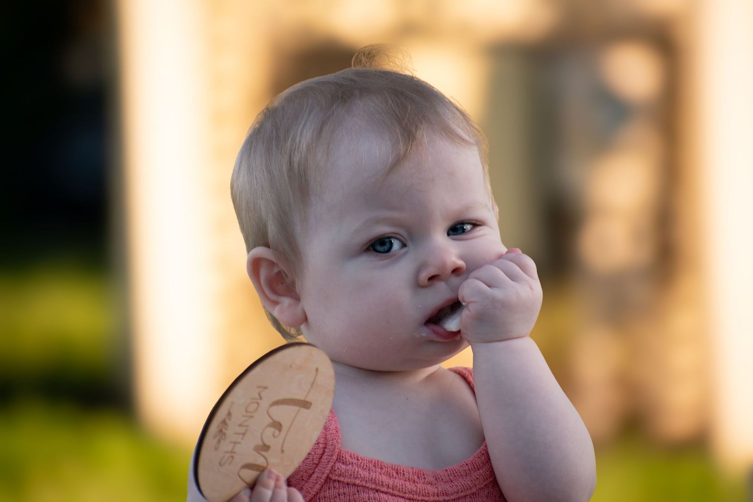 Adorable blonde baby girl in pink dress holding engraved wooden disc while putting finger in mouth.