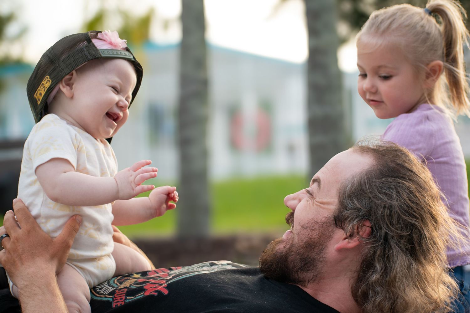 Bearded dad holding laughing baby girl while blonde toddler watches joyfully outdoors.