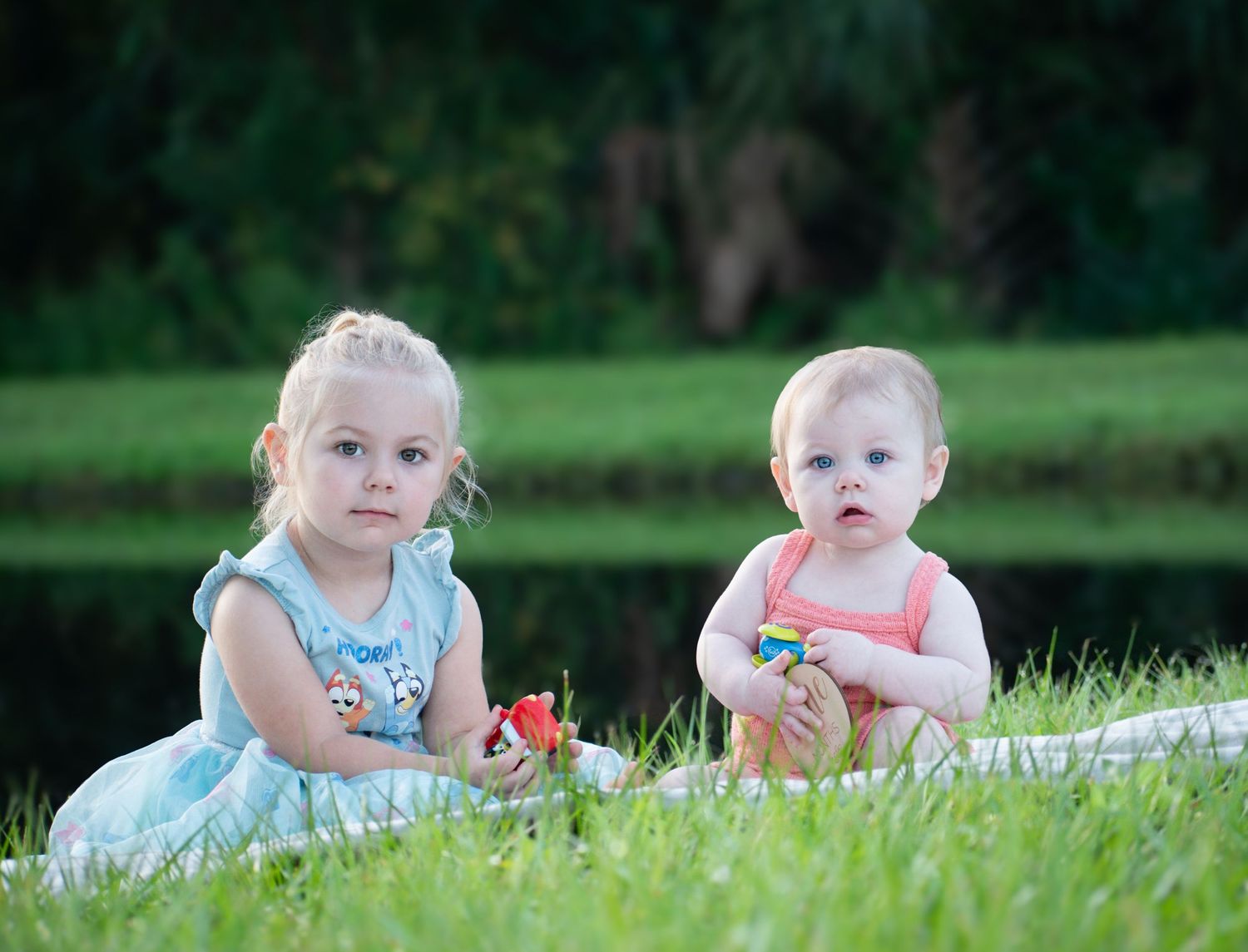 Two young sisters sit on grass outdoors, the older girl in a blue dress beside her baby sister in pink.