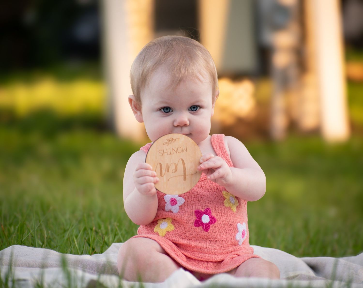 A cute baby girl in a pink floral romper chews on a wooden milestone disc in sunny grass.