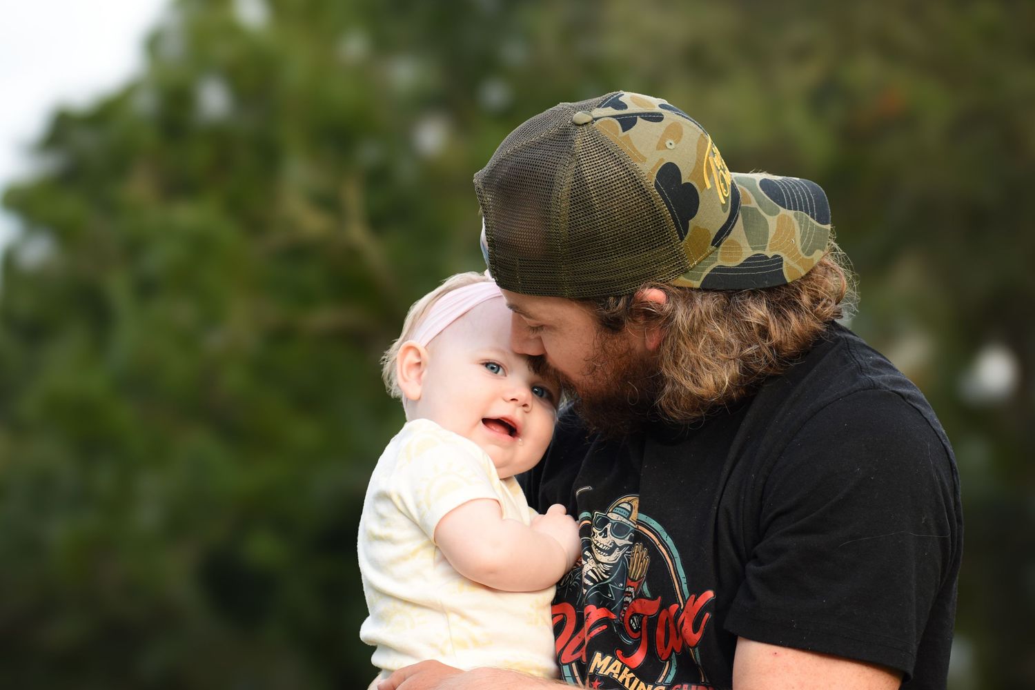 A bearded man wearing a camo hat tenderly touches foreheads with a smiling baby girl outdoors.