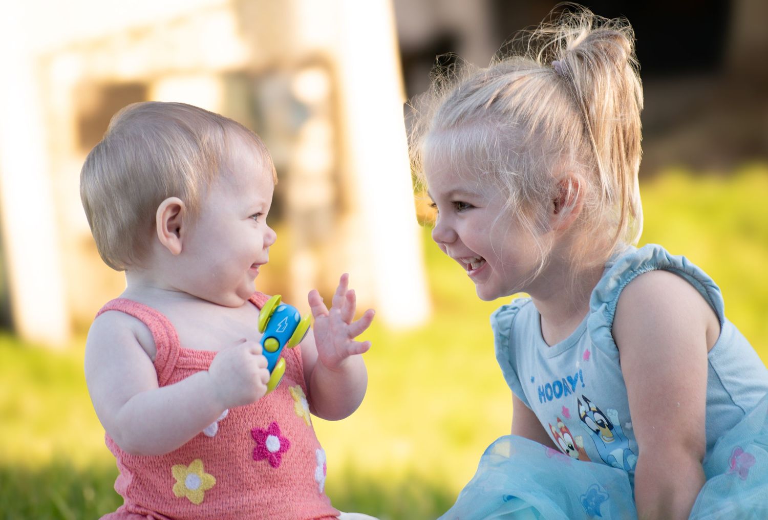 Baby in floral pink romper and smiling toddler in blue dress interact playfully on sunny green grass.